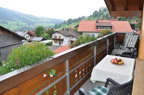 A cozy balcony adorned with flowers overlooks a quaint village nestled in lush green hills, perfect for travelers seeking a serene retreat with stunning mountain views.