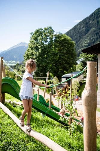 A child enjoys balancing on a wooden beam in a vibrant outdoor playground surrounded by lush greenery and towering mountains, ideal for family-friendly adventure trips.