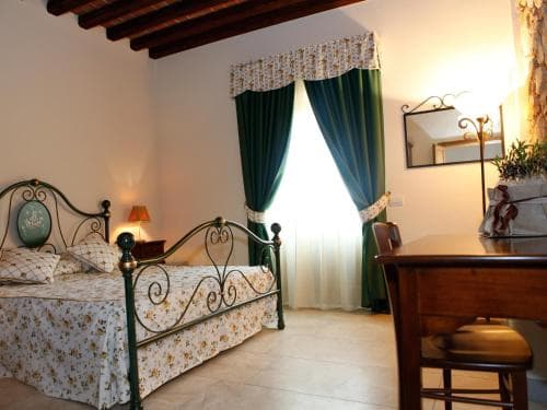 A cozy bedroom with a floral-patterned bedspread, green curtains, and warm lighting creates an inviting atmosphere, perfect for travelers seeking a comfortable retreat in a rustic villa setting.