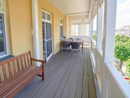 A cozy wooden balcony with a bench, dining table, and chairs offers a serene retreat, perfect for travelers seeking relaxation in a charming seaside apartment setting.