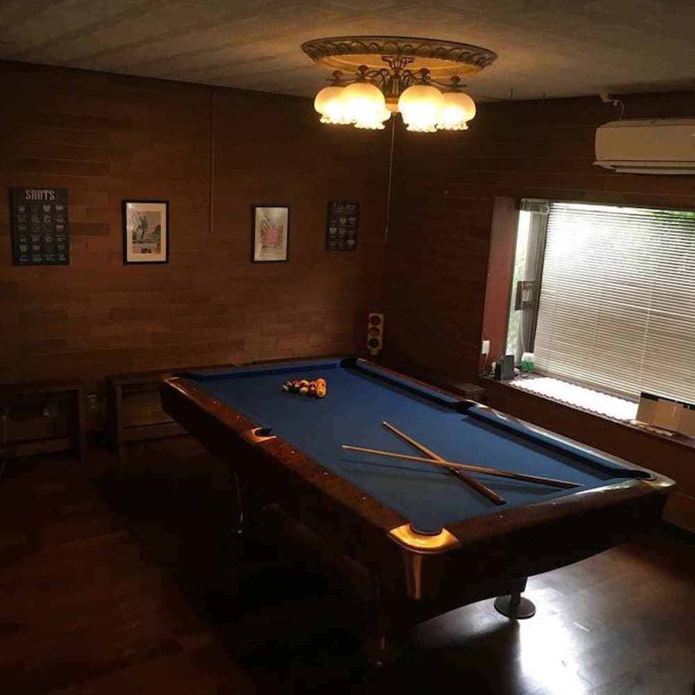 A cozy game room with a blue pool table illuminated by a vintage chandelier, wooden panel walls adorned with framed art, and a window with blinds, perfect for travelers seeking relaxation and entertainment in a rustic setting.