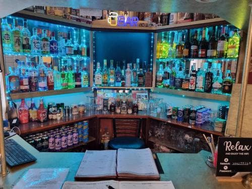A cozy bar corner illuminated by vibrant blue lights showcases an array of colorful liquor bottles, creating a relaxed and inviting atmosphere perfect for travelers seeking a unique spot to unwind and enjoy a drink.
