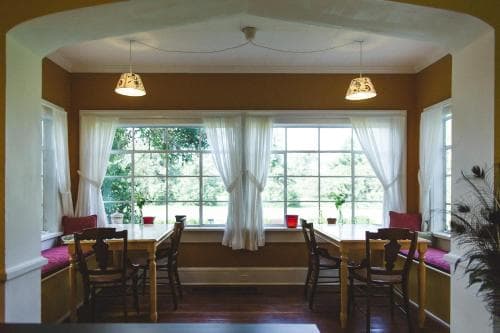 A cozy dining area with wooden tables and chairs bathed in natural light from large windows framed by white curtains, ideal for travelers seeking a serene and inviting space for meals or relaxation.