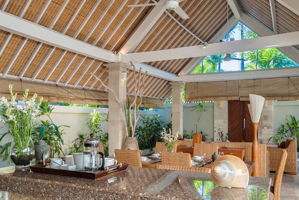 A serene tropical dining area with a thatched roof, lush greenery, and cozy wicker furniture invites travelers to relax and enjoy a tranquil meal in a resort setting.