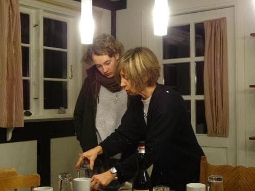 Two individuals share a cozy moment in a warmly lit dining room, pouring drinks amidst wooden furniture and soft curtains, ideal for capturing the essence of intimate gatherings and homey travel experiences.