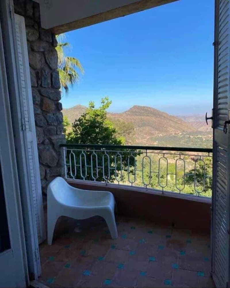A serene balcony with a white plastic chair overlooks lush green hills under a clear blue sky, ideal for travelers seeking a peaceful retreat in a mountainous region.