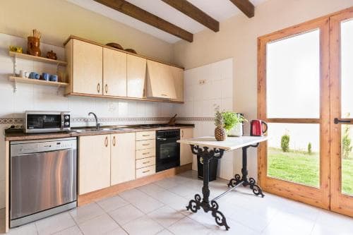 A cozy, well-lit kitchen with wooden beams, modern appliances, and a small table by the glass door leading to a green garden, ideal for travelers seeking a comfortable self-catering stay.