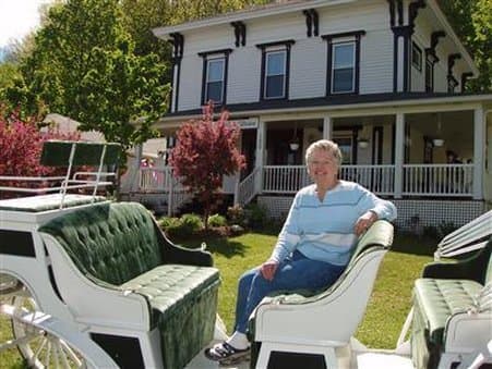 A serene scene features a smiling woman seated on a white carriage in front of a charming Victorian-style house, surrounded by lush greenery and blooming trees, ideal for a relaxing countryside getaway.
