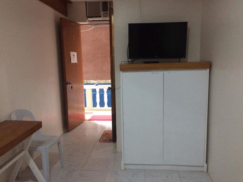 A cozy apartment interior features a simple dining setup, a white cabinet with a mounted TV, and an open door leading to a sunny balcony, ideal for travelers seeking comfort in a compact space.