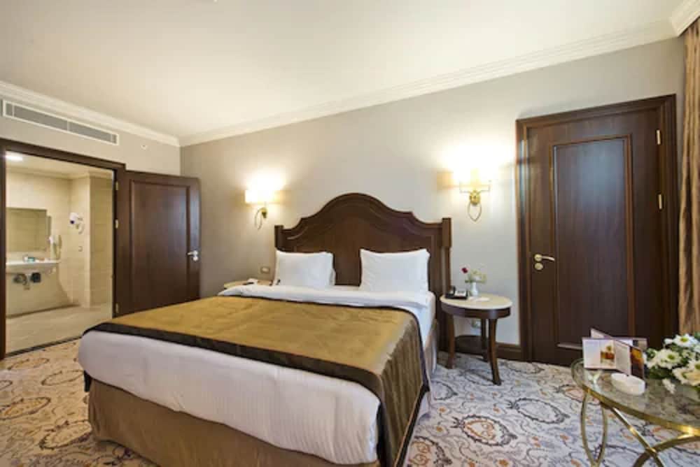 A cozy hotel room features a neatly made bed with a wooden headboard, warm lighting from wall sconces, a patterned carpet, and a glass coffee table with flowers, ideal for travelers seeking comfort and relaxation.