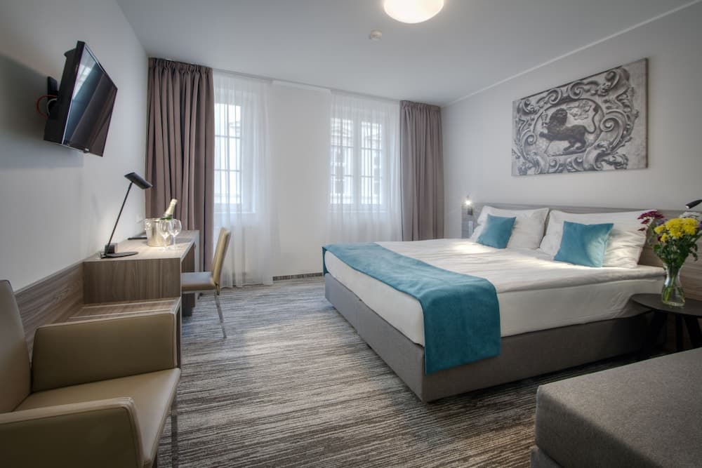 A cozy hotel room features a neatly made bed with teal accents, a desk with a chair, a wall-mounted TV, and soft natural light filtering through sheer curtains, ideal for travelers seeking comfort and relaxation.