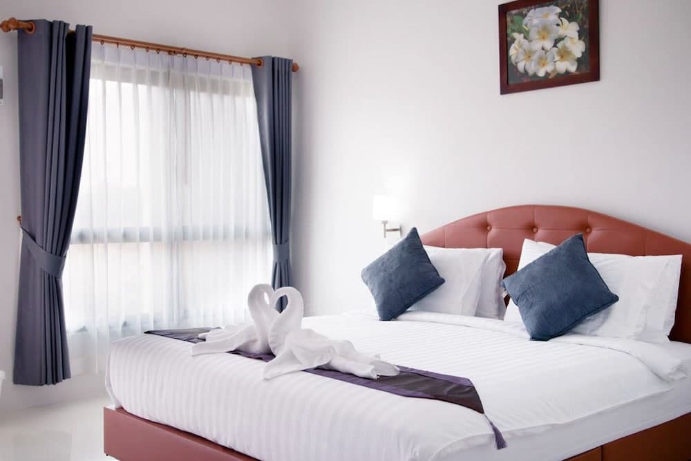 A cozy hotel room features a neatly made bed adorned with blue pillows and towel swans, framed by sheer curtains and a floral artwork, ideal for travelers seeking comfort and relaxation.