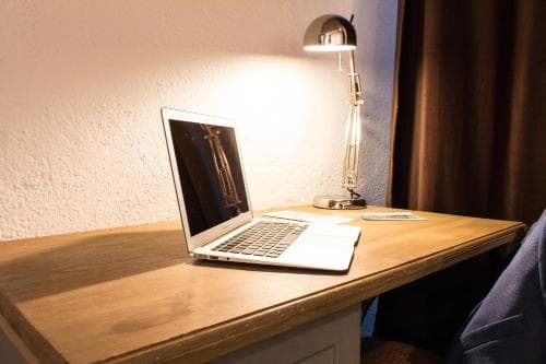 A cozy hotel room workspace features a wooden desk with an open laptop illuminated by a modern desk lamp, ideal for travelers needing a quiet spot to work or study.
