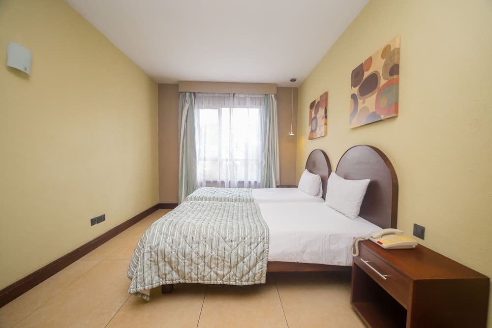 A cozy hotel room with soft yellow walls, abstract art, a neatly made bed by a bright window, and a wooden desk with a phone, ideal for travelers seeking comfort and simplicity.