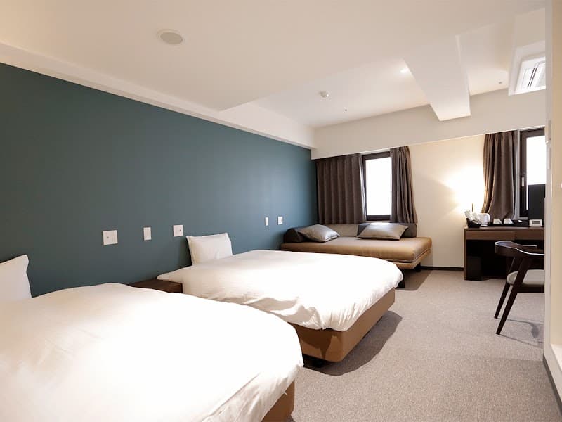 A cozy hotel room in Osaka features neatly arranged beds against a teal wall, a desk by the window, and a warm ambiance ideal for travelers seeking comfort and relaxation.