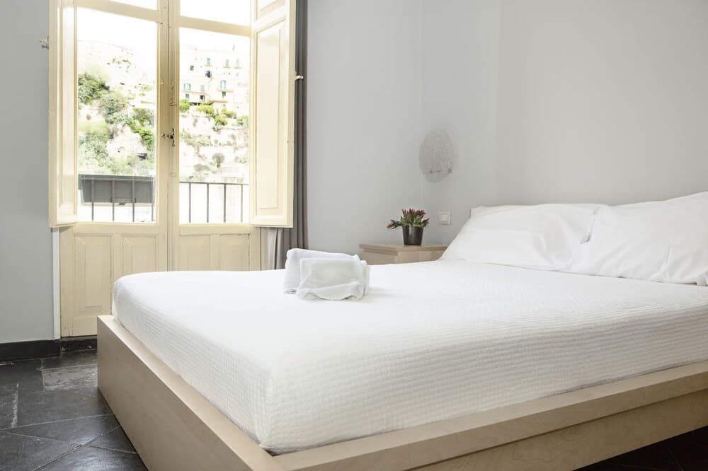 A serene minimalist bedroom with a white bed, open window revealing a scenic hillside view, and simple decor invites travelers seeking peaceful accommodation in a picturesque location.