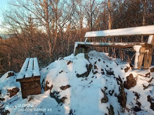 A serene winter scene captures snow-covered rocks and wooden benches amidst bare trees in a tranquil forest setting, ideal for nature photography and peaceful retreats.