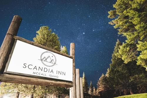 Under a starry night sky, the rustic wooden sign of Scandia Inn in McCall, Idaho stands amidst towering pine trees, evoking a serene and inviting atmosphere perfect for travelers seeking a tranquil retreat in nature.
