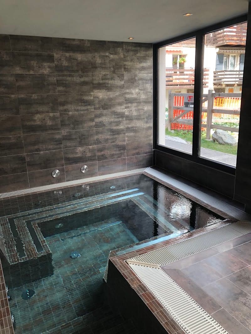 A serene indoor spa pool with modern tiled walls and a large window offering a view of quaint wooden balconies outside, ideal for relaxation at a luxury hotel in Zermatt, Switzerland.