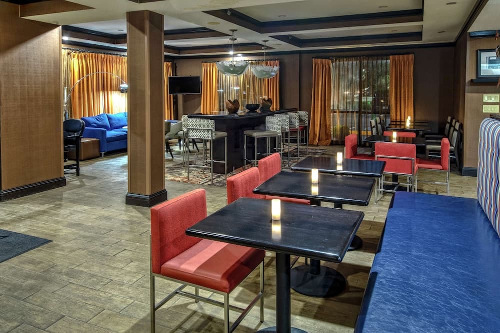 A cozy hotel lounge features vibrant red chairs, sleek black tables with candles, plush blue seating, and warm orange curtains, creating an inviting atmosphere ideal for travelers seeking relaxation or casual dining.