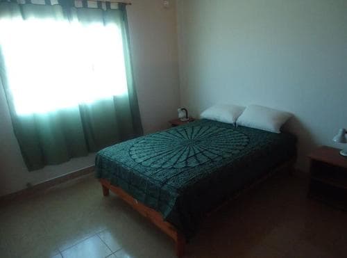 A cozy bedroom with a neatly made bed adorned in a green patterned cover, soft natural light filtering through sheer curtains, and simple bedside tables, ideal for travelers seeking a tranquil and budget-friendly accommodation.