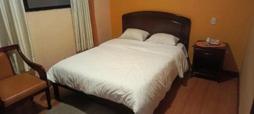 A cozy hotel room features a neatly made bed beside a wooden chair and nightstand, set against warm yellow walls, ideal for travelers seeking comfort and simplicity.