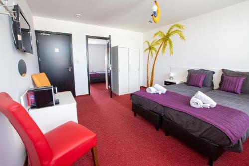 A cozy hotel room with vibrant red carpeting, tropical-themed decor including palm tree wallpaper, a comfortable bed with purple linens, and modern amenities like a flat-screen TV, ideal for travelers seeking a relaxing and stylish stay.