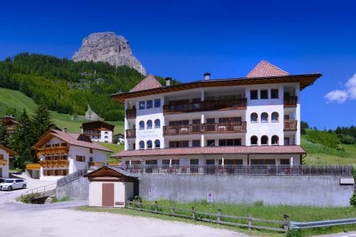 A charming alpine hotel with balconies overlooks a lush green valley under a clear blue sky, ideal for travelers seeking a serene mountain retreat.