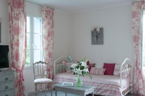 A cozy bedroom with pink toile curtains, a white metal bed adorned with striped bedding, and a vase of fresh flowers on a table, evokes a serene and charming atmosphere ideal for a relaxing getaway.