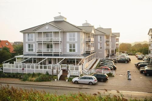A serene coastal town features a charming multi-story apartment complex with balconies, surrounded by parked cars and lush greenery, ideal for travelers seeking a peaceful seaside retreat.