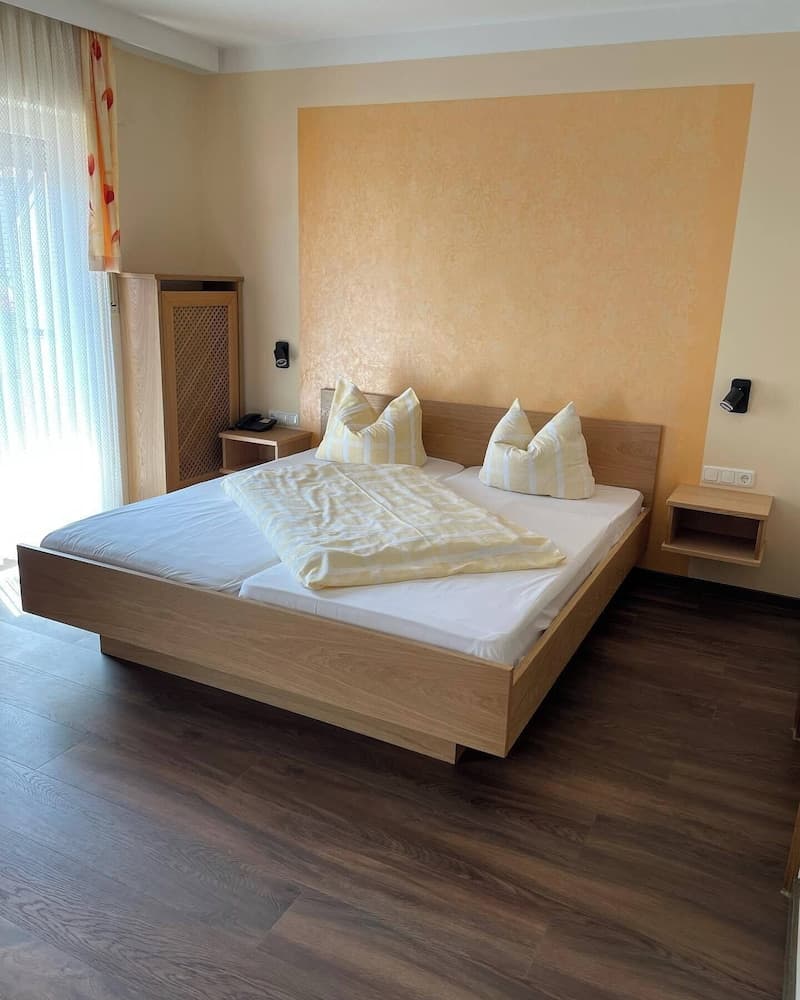 A cozy hotel room features a neatly made bed with yellow bedding against a warm-toned wall, wooden flooring, and natural light streaming through sheer curtains, ideal for travelers seeking comfort and relaxation.