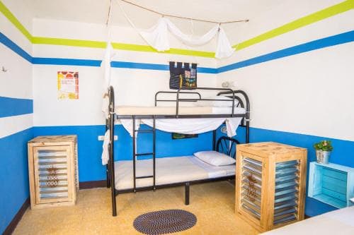 A cozy hostel room with vibrant blue and green striped walls, bunk beds adorned with white drapes, wooden storage crates, and a relaxed atmosphere ideal for budget travelers seeking comfort.