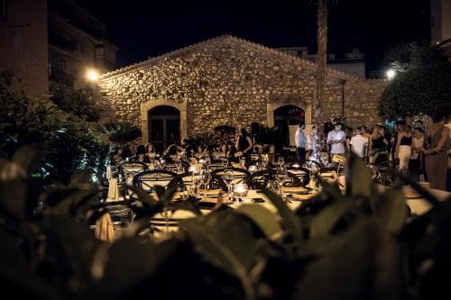 A cozy outdoor evening gathering unfolds in front of a rustic stone building, illuminated by warm lights, surrounded by lush greenery, perfect for intimate dinner parties or cultural events.