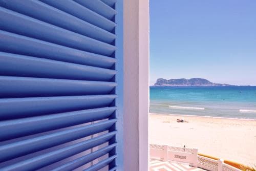A serene coastal view framed by blue shutters invites travelers to relax on the sandy beach under clear skies, ideal for vacation planning and destination inspiration.