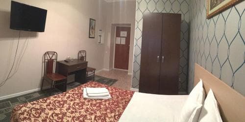 A cozy hotel room features a neatly made bed with patterned bedding, a wooden desk with chairs, a wardrobe, and a wall-mounted TV, ideal for travelers seeking comfort and functionality in a compact space.