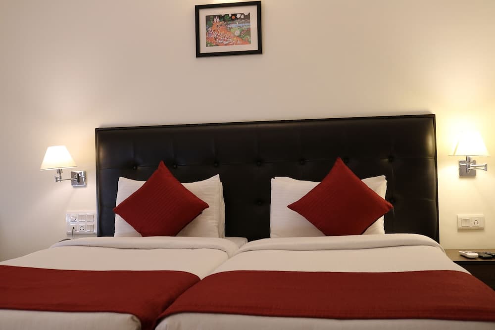 A cozy hotel room features two neatly arranged beds with red accents, a black leather headboard, wall-mounted lamps, and a framed artwork, ideal for travelers seeking comfort and relaxation.