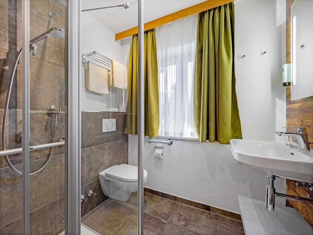 A modern bathroom with a glass-enclosed shower, sleek white fixtures, green curtains, and wooden accents offers a clean and inviting space ideal for travelers seeking comfort and style.
