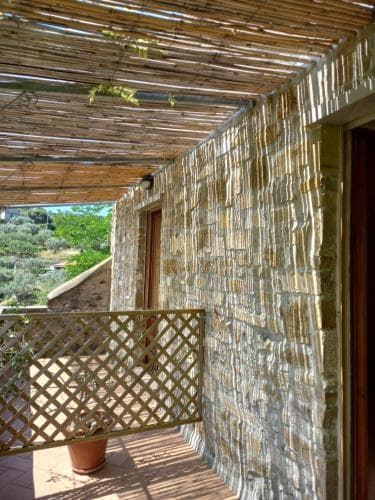A serene stone-walled balcony shaded by a rustic bamboo roof overlooks lush greenery, ideal for travelers seeking a tranquil retreat in a countryside villa.