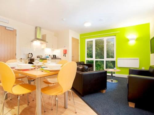 A cozy apartment features a vibrant lime green accent wall, modern furniture including yellow chairs and a wooden dining table, set against large windows with lush greenery outside, ideal for travelers seeking a stylish and comfortable stay.