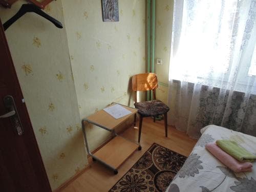 A cozy, modestly furnished bedroom with floral wallpaper, a wooden chair, and soft natural light filtering through lace curtains, ideal for budget travelers seeking simple accommodations.