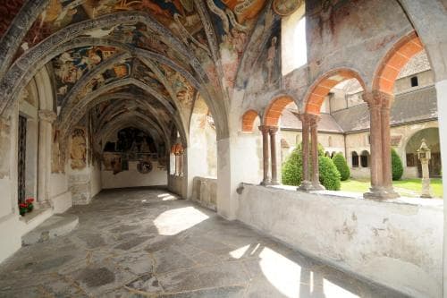 A serene monastery cloister features arched walkways adorned with vibrant frescoes, stone columns, and lush greenery, ideal for travelers seeking historical and spiritual exploration.
