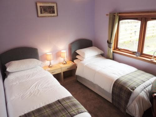 A cozy guesthouse room with two neatly made single beds adorned with plaid blankets, soft purple walls, warm bedside lamps, and a window offering a serene outdoor view, ideal for travelers seeking a peaceful retreat.