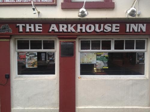 The Arkhouse Inn, a cozy pub with red accents and window posters, invites travelers seeking a warm atmosphere and local charm for a relaxing stopover.