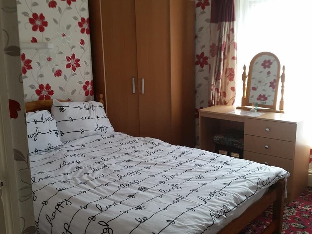 A cozy bedroom with floral wallpaper and curtains, featuring a wooden bed with a "love sleep happy" duvet, a wooden wardrobe, and a dressing table with a mirror, ideal for travelers seeking a comfortable and homey accommodation.