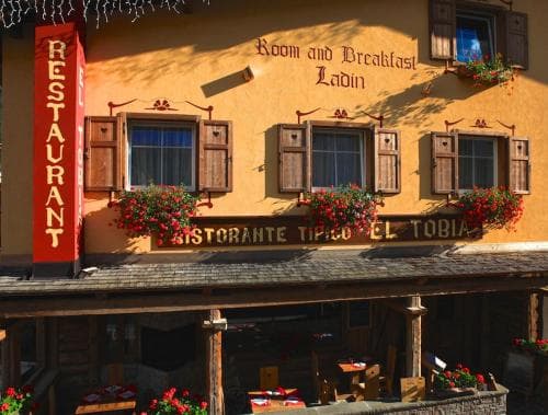 A charming yellow building adorned with wooden shutters and vibrant flower boxes offers a cozy "Room and Breakfast Ladin" experience, perfect for travelers seeking a quaint European getaway.