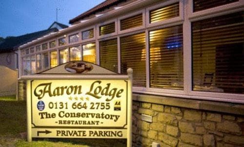 A cozy evening view of Aaron Lodge, a charming conservatory restaurant with warm interior lighting, stone exterior, and a welcoming signboard, ideal for travelers seeking a comfortable dining experience with private parking.