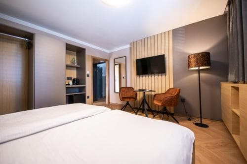 A cozy hotel room features a neatly made bed, modern furnishings including leather chairs and a sleek desk, warm lighting from a patterned floor lamp, and a minimalist design ideal for travelers seeking comfort and style.