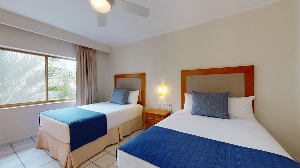 A cozy hotel room with two neatly made beds adorned with blue accents, a warm bedside lamp, and a window view of lush palm trees, ideal for travelers seeking comfort and relaxation.