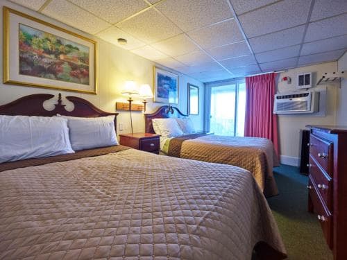 A cozy hotel room features two neatly made beds under warm lighting, framed artwork on the walls, and a view of a balcony through a window, ideal for travelers seeking comfort and relaxation.
