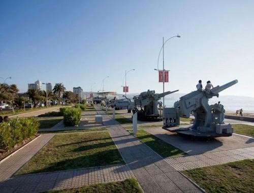 A serene seaside promenade features historical artillery pieces on display, inviting travelers to explore its blend of military heritage and coastal tranquility under clear skies.